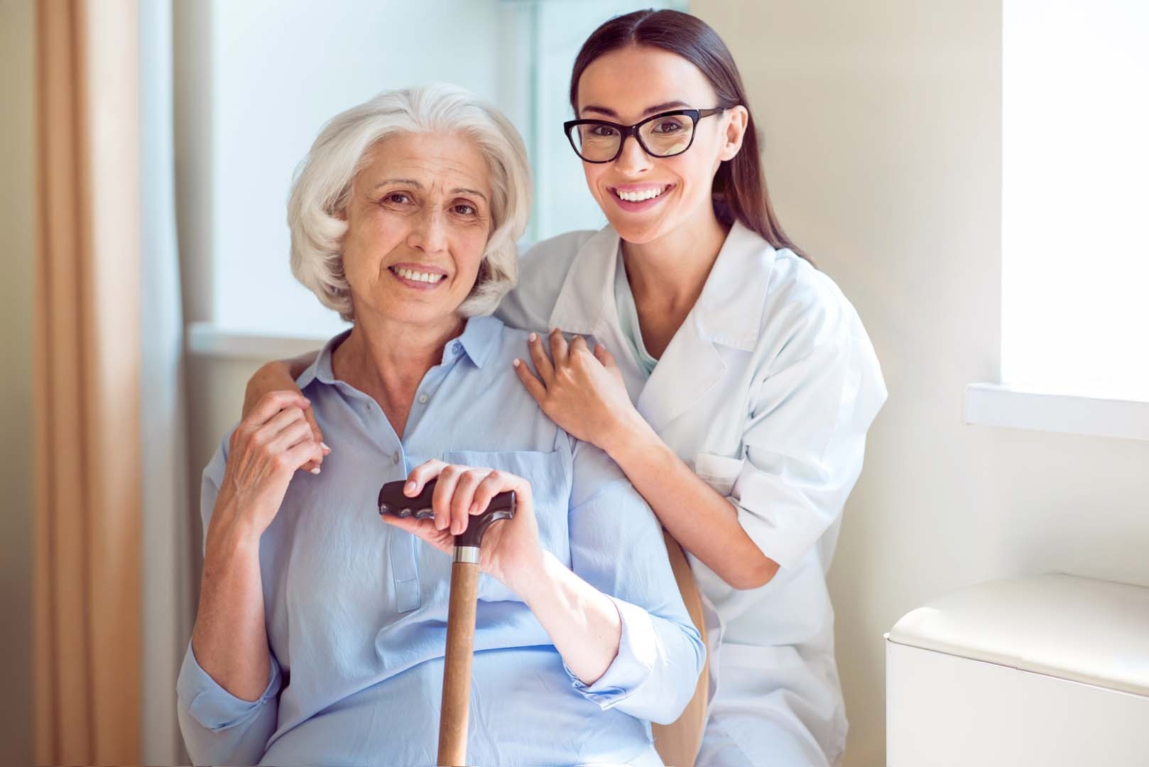 Smiling elderly woman with a cane sits beside a caregiver in a white coat, who rests a hand on her shoulder in a bright room.