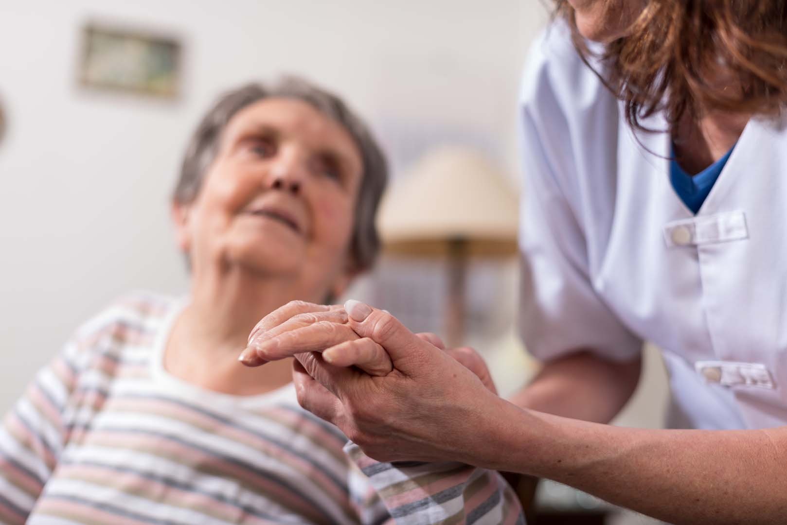 Nurse in white uniform holds an elderly patient's hands in a comforting caregiving gesture.