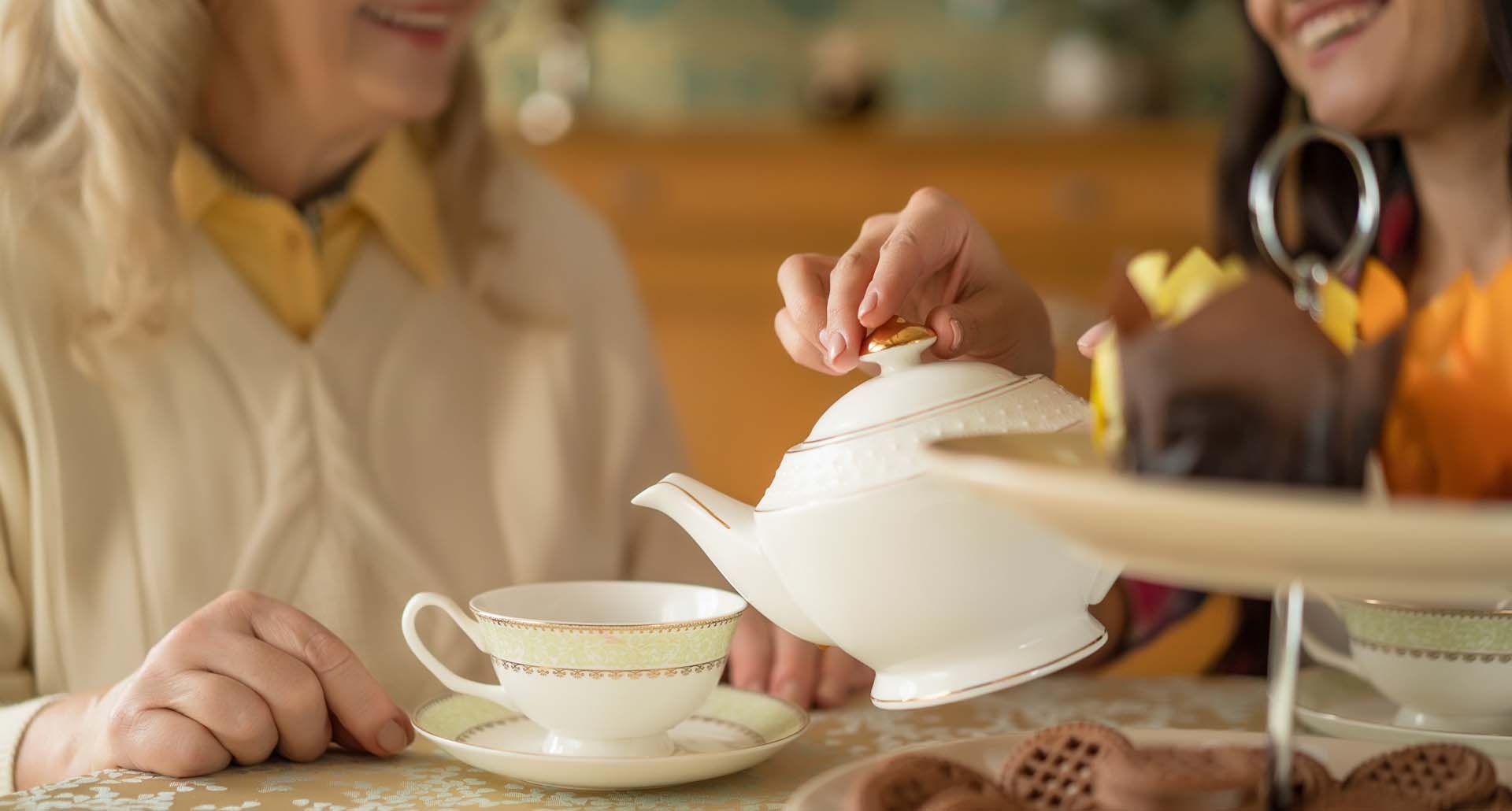 Two people share tea as one pours from a white teapot into a patterned teacup, with cookies on the table nearby.