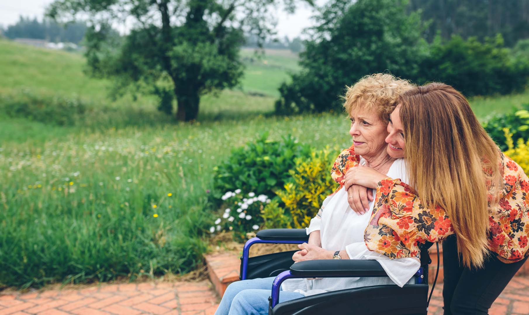 Two women in wheelchairs outdoors in a sunny garden, one supporting the other with a warm embrace from behind.