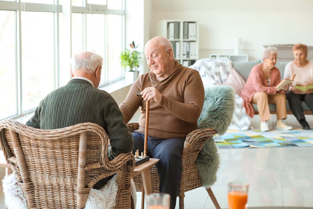 Senior men playing chess in nursing home - Calm Seas Home Care Two elderly men sit in wicker chairs by a sunny window, playing chess with a cane on a small table between them, while others read on a couch in the background.