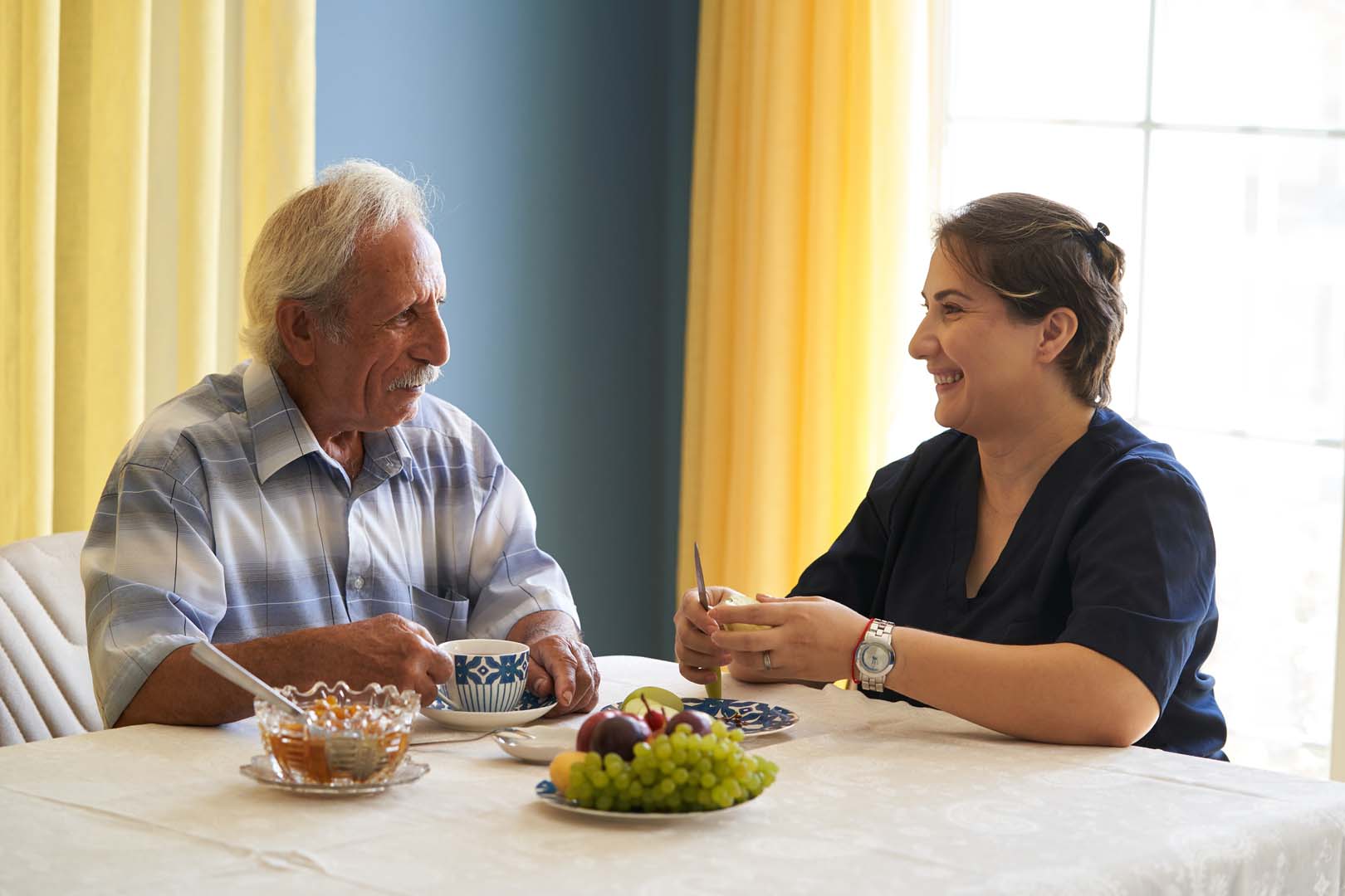 Older man and a female caregiver sharing tea at a sunny dining table with fruit in the foreground.