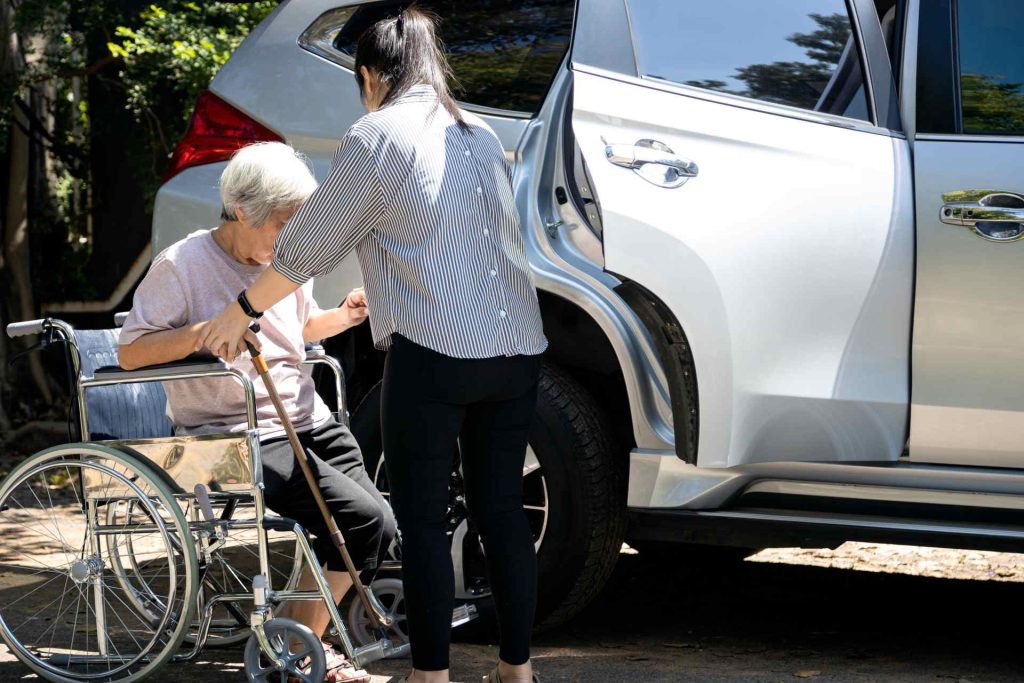 Calm Seas Home Care - Calm Seas Home Care Caregiver helping an elderly woman in a wheelchair prepare to enter a silver SUV with its door open.