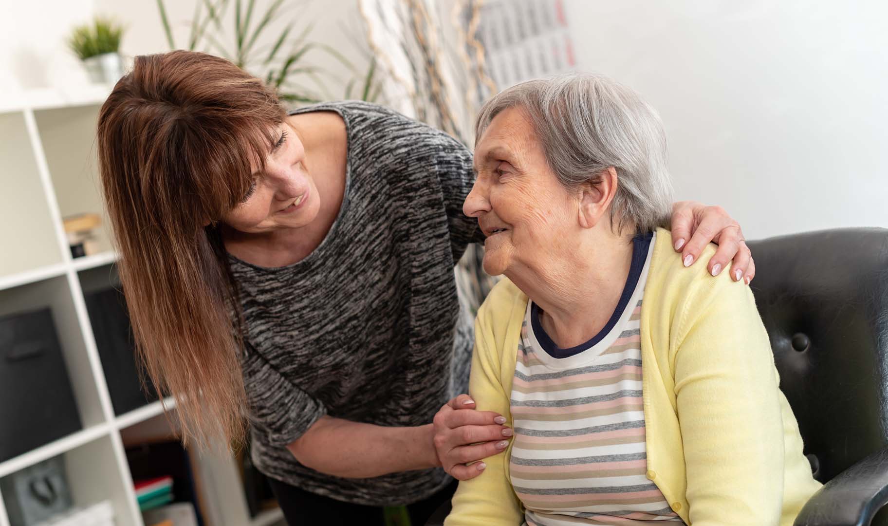 Caregiver leans in with a smile as she comforts an elderly woman sitting on a sofa, hand resting on her shoulder in a living room light.