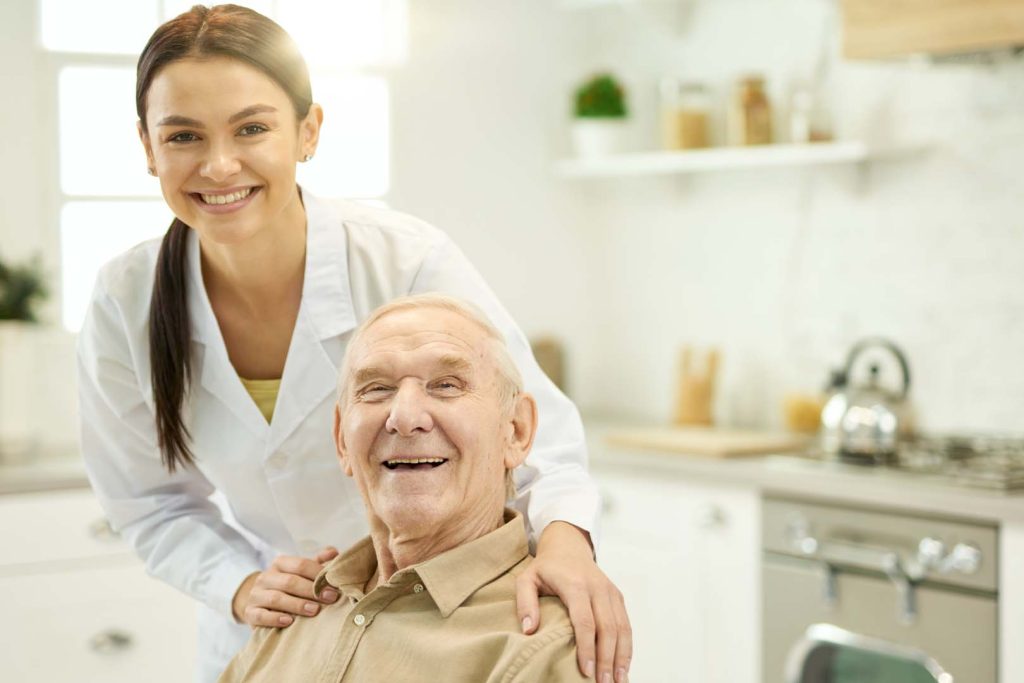 Pleased aged man being visited by a qualified doctor at home - Calm Seas Home Care Smiling healthcare worker assists an elderly man in a bright home kitchen in a moment of care and companionship.