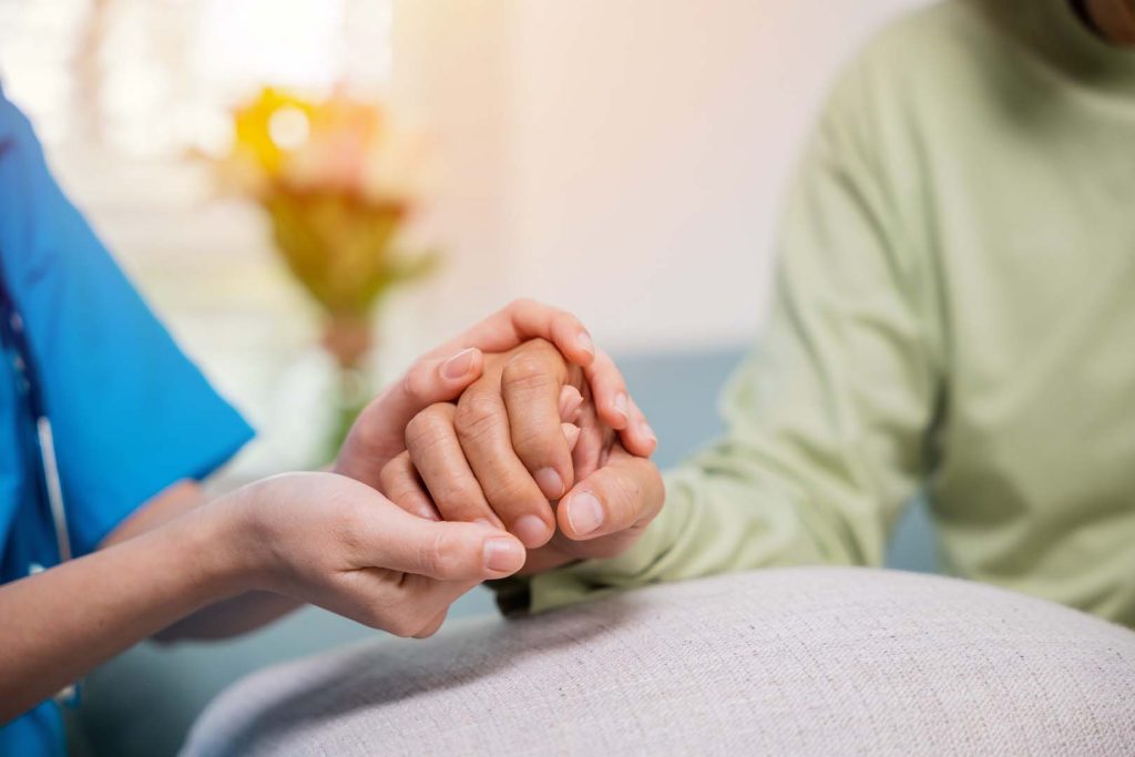 Young woman helping hold hands offering her senior man - Calm Seas Home Care Healthcare professional gently holds a patient's hands in a comforting gesture.
