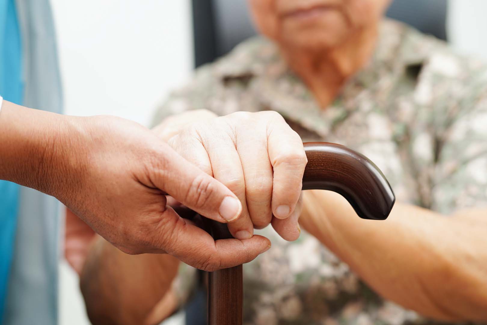 Two hands firmly grasp a dark wooden cane, depicting assistance to an elderly person in the background.]Another option: Close-up of a caregiver helping an elderly person hold a cane during support.