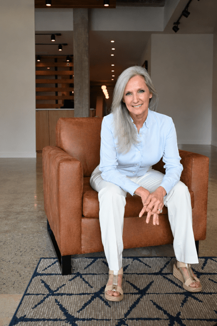 Valerie Hymel - Calm Seas Home Care Woman with gray hair sits on a brown leather armchair in a modern lobby, smiling at the camera wearing a light blue shirt and white pants.